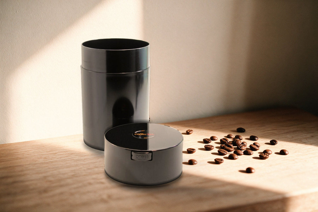 Il contenitore sottovuoto CoffeeVac nero aperto sul piano di legno, con il coperchio appoggiato accanto e i chicchi sparsi