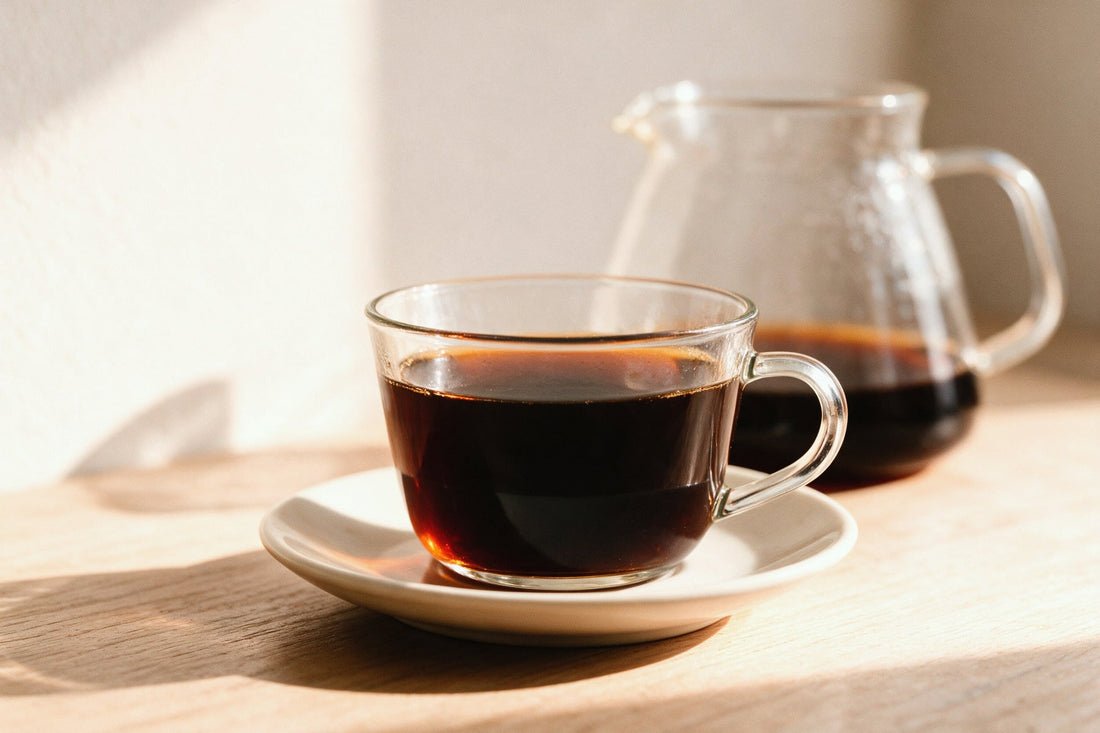 Una tazza di vetro con caffe' filtro appena versato, sul piattino, e la caraffa dell'estrazione sullo sfondo
